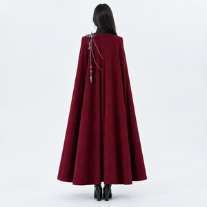 May include: A floor-length, deep red cape with a high collar, worn over a black garment. The cape features a silver-toned metal accessory on the right shoulder, consisting of a decorative emblem with dangling chains and a pointed, spear-like charm.