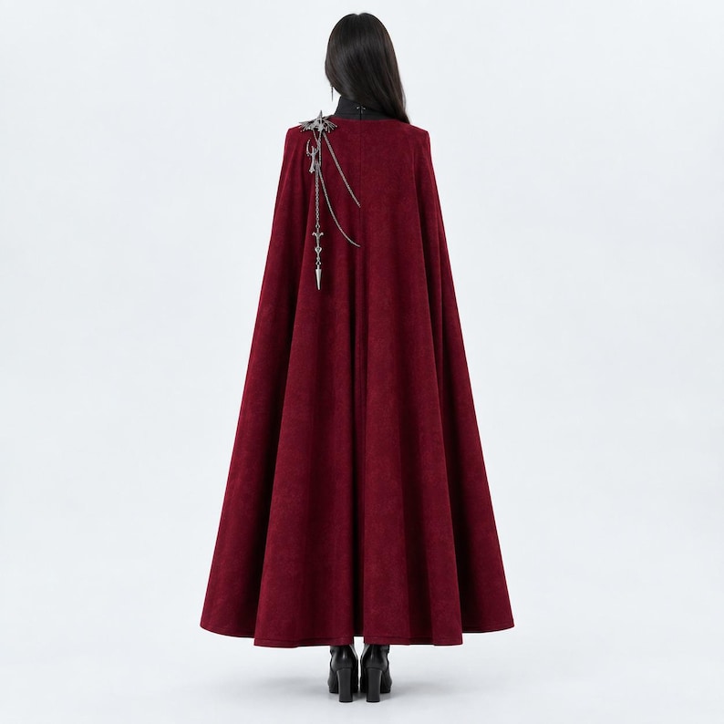 May include: A floor-length, deep red cape with a high collar, worn over a black garment. The cape features a silver-toned metal accessory on the right shoulder, consisting of a decorative emblem with dangling chains and a pointed, spear-like charm.