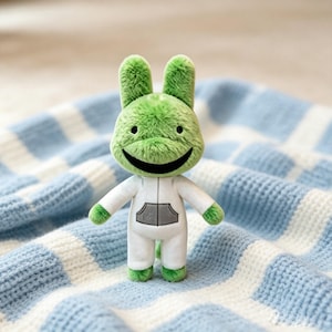 May include: A plush toy featuring a green, fuzzy, rabbit-like creature with long ears and a wide, black, stitched smile. The figure wears a white, long-sleeved jumpsuit with a gray, rectangular front pocket detail, and has green hands and feet.