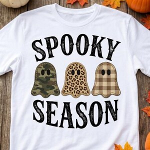 May include: A white t-shirt featuring the black text SPOOKY SEASON in a distressed font. Between the words are three ghost-shaped graphics: one with a green camouflage pattern, one with a brown leopard print, and one with a brown plaid pattern.