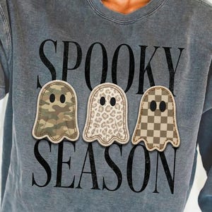 May include: A charcoal gray acid-wash sweatshirt featuring the black text SPOOKY SEASON. Three ghost-shaped patches are centered between the words, each with black oval eyes and a rope-style border. The ghosts feature camouflage, leopard, and checkerboard patterns.