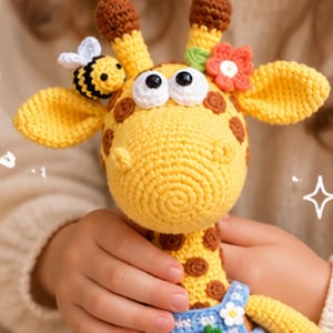 May include: A yellow and brown crocheted giraffe toy featuring a small crocheted bee on one ear and a red flower with a green leaf on the other. The giraffe has large white eyes with black pupils and wears blue overalls with a small white flower detail.