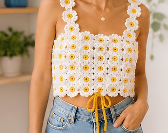 Puede incluir: Top de crochet blanco con detalles de margaritas amarillas. El top presenta un escote cuadrado y tirantes, con un cordón amarillo en la cintura. El patrón de margaritas se repite en toda la prenda.