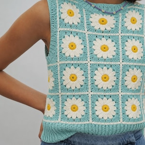May include: A sleeveless crochet top featuring a grid pattern of light blue squares, each containing a white daisy with a yellow centre. The garment is constructed from individual floral motifs joined by white yarn, finished with a ribbed hem.
