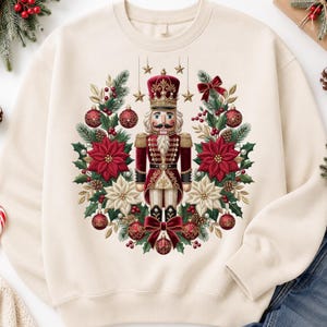 May include: A cream-colored crewneck sweatshirt featuring a central graphic of a nutcracker soldier surrounded by a festive wreath. The wreath includes red and white poinsettias, pine branches, red ornaments, pinecones, and gold hanging stars.