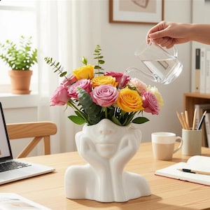 May include: A white ceramic face vase filled with pink, yellow, and orange roses. A person is watering the flowers with a clear glass watering can. The vase sits on a wooden desk. The flowers are a vibrant display.