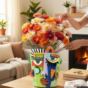 Op de afbeelding: Een kleurrijke, abstracte gezichtsvormige vaas bevat een levendig boeket van oranje, rode en gele bloemen. De vaas heeft een groene basis met blauwe, gele en rode gelaatstrekken. De bloemen worden water gegeven.