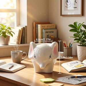 Puede incluir: Una maceta blanca de cerámica con forma de elefante, de 6,6 cm de ancho y 7 cm de alto, con orejas internas rosas y ojos pintados. La maceta está sobre un escritorio de madera con un cuaderno, un bolígrafo y notas adhesivas.