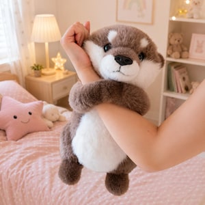 Puede incluir: Un juguete de peluche de nutria marrón y blanco es sostenido. El juguete tiene una barriga blanca, patas marrones oscuras y una nariz negra. El animal de peluche está diseñado para parecerse a una nutria real.