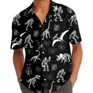 May include: A black short-sleeve button-down shirt featuring a repeating white graphic print of Bigfoot figures, various dinosaur skeletons, and spider webs. The shirt has a classic collar and a front button closure with white buttons.