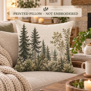 Puede incluir: Un cojín cuadrado con un paisaje forestal impreso que muestra altos árboles de hoja perenne y follaje denso en tonos verdes, marrones y crema. El texto PRINTED PILLOW - NOT EMBROIDERED aparece en la parte superior de la imagen.
