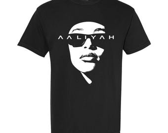 May include: A black short-sleeve t-shirt featuring a high-contrast white graphic print of a face wearing sunglasses. The text AALIYAH is printed in a bold, stylized white font across the center of the sunglasses and the upper face area.
