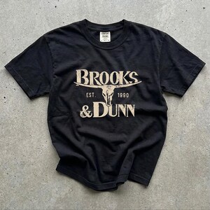 May include: A black short-sleeve t-shirt featuring a beige graphic print. The design displays the text BROOKS & DUNN with a steer skull illustration in the center and the text EST. 1990 below the horns. The shirt has a crew neckline.