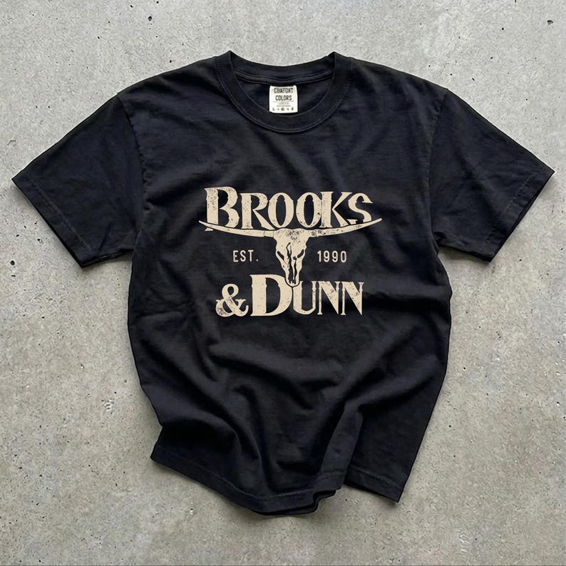 May include: A black short-sleeve t-shirt featuring a beige graphic print. The design displays the text BROOKS & DUNN with a steer skull illustration in the center and the text EST. 1990 below the horns. The shirt has a crew neckline.