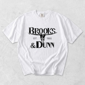 May include: A white short-sleeved t-shirt featuring a black graphic print on the chest. The design includes the text BROOKS & DUNN, the phrase EST. 1990, and a central illustration of a longhorn steer skull with a distressed, weathered texture.