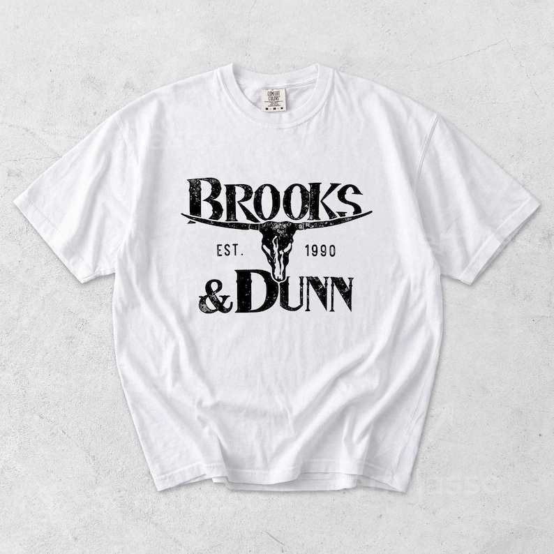 May include: A white short-sleeved t-shirt featuring a black graphic print on the chest. The design includes the text BROOKS & DUNN, the phrase EST. 1990, and a central illustration of a longhorn steer skull with a distressed, weathered texture.