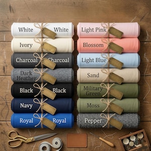 May include: A collection of rolled fabric bolts in various colors including white, ivory, charcoal, dark heather, black, navy, royal, light pink, blossom, light blue, sand, military green, moss, and pepper, each tied with twine and a blank tag.