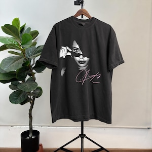 May include: Black t-shirt featuring a grayscale image of a person with sunglasses, with a pink signature below. The t-shirt is hanging on a wooden hanger against a white wall. A tall green plant is to the left.