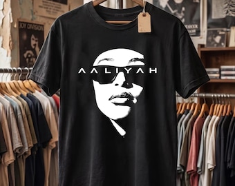 May include: A black short-sleeve t-shirt featuring a high-contrast white graphic of a face wearing sunglasses. The name AALIYAH is printed in white capital letters across the center of the sunglasses. A blank brown rectangular tag hangs from the collar.