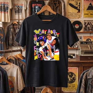 May include: A black short-sleeve t-shirt featuring a graphic print of three individuals and the colourful text "WHAT ABOUT your FRIENDS". The design includes a person wearing a patterned head covering, a person in a white shirt, and a person in yellow.