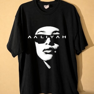 May include: Black t-shirt featuring a black and white graphic of a person wearing sunglasses. The word "AALIYAH" is printed in white above the graphic. The shirt has short sleeves and a crew neck.