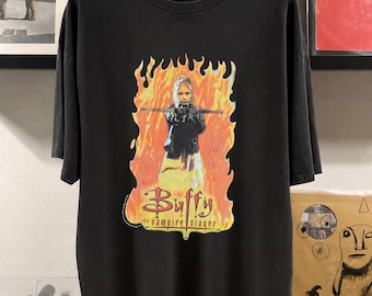 May include: Black t-shirt featuring a graphic of a woman holding a stake, set against a fiery background. The text "Buffy the Vampire Slayer" is displayed below the image in a stylized font. The shirt has short sleeves.