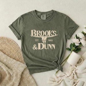 May include: An olive green short-sleeve t-shirt featuring a cream-colored graphic of a longhorn skull centered above the text "BROOKS & DUNN" and "EST. 1990". The shirt has a crew neckline and a slightly distressed, vintage-inspired fabric texture.
