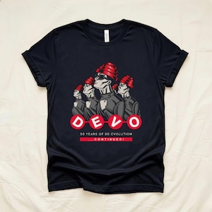 May include: A black short-sleeved t-shirt featuring a graphic of four figures wearing red energy dome hats. Below the figures, the text reads DEVO, 50 YEARS OF DE-EVOLUTION, and ...CONTINUED! in white and red lettering on the front of the shirt.