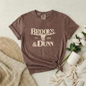 May include: A brown short-sleeved t-shirt featuring a cream-colored graphic of a steer skull centered between the text BROOKS & DUNN and EST. 1990. The shirt has a crew neckline and a tag at the inner collar indicating the brand and size.