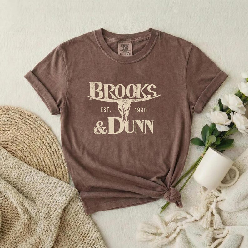 May include: A brown short-sleeved t-shirt featuring a cream-colored graphic of a steer skull centered between the text BROOKS & DUNN and EST. 1990. The shirt has a crew neckline and a tag at the inner collar indicating the brand and size.