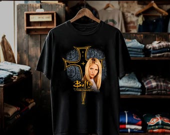 May include: A black short-sleeved t-shirt featuring a graphic print of a blonde woman centred in front of a large, ornate gold-outlined cross. The text "Buffy the Vampire Slayer" is printed in gold lettering below the cross and the woman's portrait.