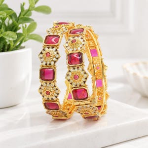 Peut inclure: Deux bracelets rigides dorés ornés d'une alternance de motifs hexagonaux et rectangulaires. Chaque pièce est incrustée de pierres en verre facettées rose foncé et de détails en émail blanc. Le métal présente une finition texturée et ornée.