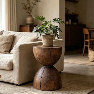 May include: A rustic, wooden side table with a unique hourglass shape. The table is made of dark brown wood and features a potted plant with green leaves on top. The table is placed next to a beige sofa in a living room setting.