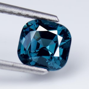 May include: A cushion-cut deep blue gemstone with a faceted surface that reflects light, held by a pair of metal tweezers. The stone displays a rich, saturated teal-blue hue with sharp geometric facets and clear internal reflections throughout.