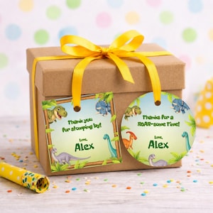 Può includere: Una scatola regalo quadrata in cartone marrone legata con un nastro di raso giallo brillante. Due etichette di carta sono attaccate al nastro: un'etichetta rettangolare con illustrazioni di dinosauri e il testo "Thank you for stomping by! Love, Alex," e un'etichetta circolare con grafiche di dinosauri e il testo "Thanks for a ROAR-some time! Love, Alex."