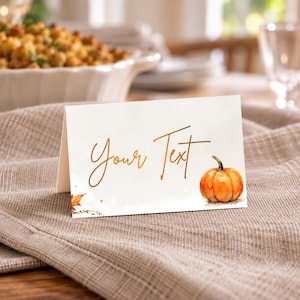 May include: A white folded place card featuring the handwritten-style text Your Text in a golden-brown color. The design includes a watercolor illustration of a small orange pumpkin on the right side and a single autumn leaf on the bottom left.