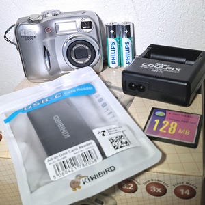 Op de afbeelding: Een zilveren Nikon Coolpix 3100 digitale camera, twee Philips AA-batterijen, een zwarte Nikon MH-70 batterijlader, een 128 MB Compact Flash-kaart en een Kiwibird USB-C kaartlezer in de originele verpakking, rustend op de cameradoos.