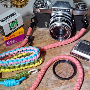 Könnte beinhalten: Eine silber-schwarze Icarex 35 Kamera mit Carl Zeiss Objektiv, befestigt an einem rosa Seilriemen. Enthalten sind eine Schachtel Ilford HP5 Plus 400 Film, eine Schachtel Kodak Gold Film, ein schwarzer Objektivfilter und ein buntes Paracord-Zubehör.