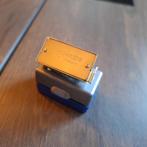 Könnte beinhalten: Ein kleiner, rechteckiger Tacker in Grau und Blau mit einer goldfarbenen Metallplatte oben. Auf der Platte steht "Auto Knips" und "Made in Germany". Der Tacker liegt auf einer Holzoberfläche.