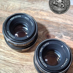 Könnte beinhalten: Zwei schwarze Pentacon auto 1.8/50 Kameraobjektive mit silbernen Metallakzenten und Glaselementen. Die Objektive weisen weiße Beschriftungen auf, darunter der Markenname, die Blende und die Seriennummern 5987917 und 6833701 auf den Frontringen.
