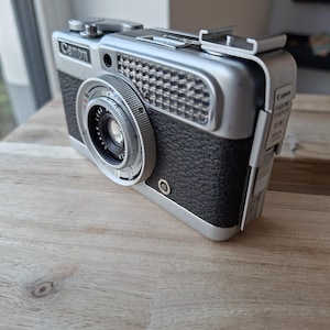 May include: A silver and black Canon Demi film camera featuring a textured grip, a circular lens with manual focus rings, a rectangular grid light meter window, and a metal flash unit coupler attached to the top. The side displays Canon branding.