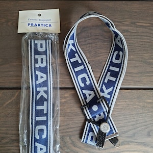 Puede incluir: Una correa de cámara Praktica azul y blanca con la palabra "PRAKTICA" repetida. La correa está empaquetada en plástico transparente y también se muestra ensamblada. La correa tiene un cierre de cuero negro y metal.