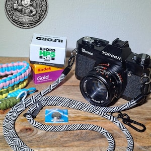 May include: A black Topcon Unirex camera with a UV Topcor 50mm lens, attached to a black and white patterned rope camera strap. Included are a box of Ilford HP5 Plus 400 film, a box of Kodak Gold film, a 1.5V battery, and two braided paracord wrist straps.