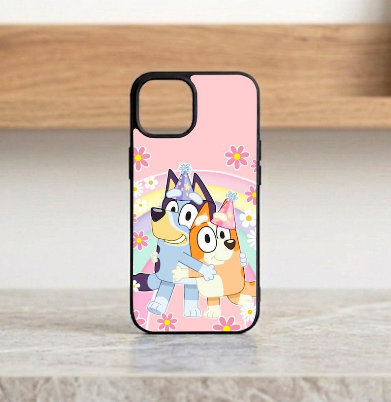 Pu&ograve; includere: Custodia per telefono rosa con un'illustrazione a cartoni animati di due cani che si abbracciano, indossano cappelli da festa e sono ambientati su uno sfondo di arcobaleno e fiori. La custodia ha un bordo nero e un ritaglio per la fotocamera.