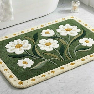 May include: A rectangular bath mat featuring a deep green center with a cluster of white flowers with yellow centers and green leaves. The mat has a cream-colored border decorated with small yellow leaf motifs along the entire perimeter.