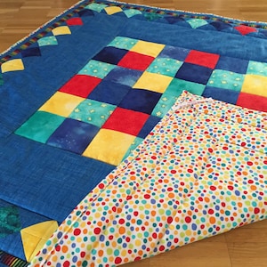 Peut inclure: Une couverture matelassée présentant une grille centrale de carrés rouges, jaunes, bleus et sarcelle avec des motifs d'étoiles. La bordure est bleu uni avec des losanges, et le coin replié révèle un envers blanc à pois multicolores.