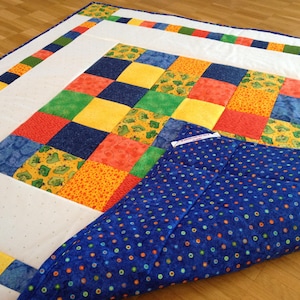 Könnte beinhalten: Eine Patchwork-Decke mit einem zentralen Raster aus bunten Quadraten in Rot, Gelb, Blau und Grün, umgeben von einem weißen Rand mit einer farbigen quadratischen Kante. Die Unterseite besteht aus blauem Stoff mit kleinen, bunten Punkten.