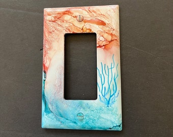 May include: A light switch cover with a blue and orange abstract design featuring a blue coral branch.