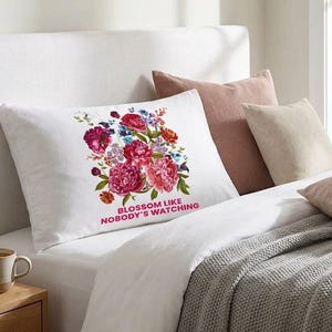 Op de afbeelding: Een witte rechthoekige kussensloop met een kleurrijk boeket van roze, rode en blauwe bloemen. Onder de bloemen staat de tekst BLOSSOM LIKE NOBODY'S WATCHING in dikke, roze hoofdletters. De kussensloop ligt op een opgemaakt bed.