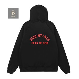 Op de afbeelding: Een zwarte hoodie met lange mouwen en de tekst ESSENTIALS FEAR OF GOD in een opvallend rood lettertype op de achterkant. Het kledingstuk heeft een capuchon, lange mouwen met geribbelde boorden en een rechte zoom in een effen zwarte kleur.